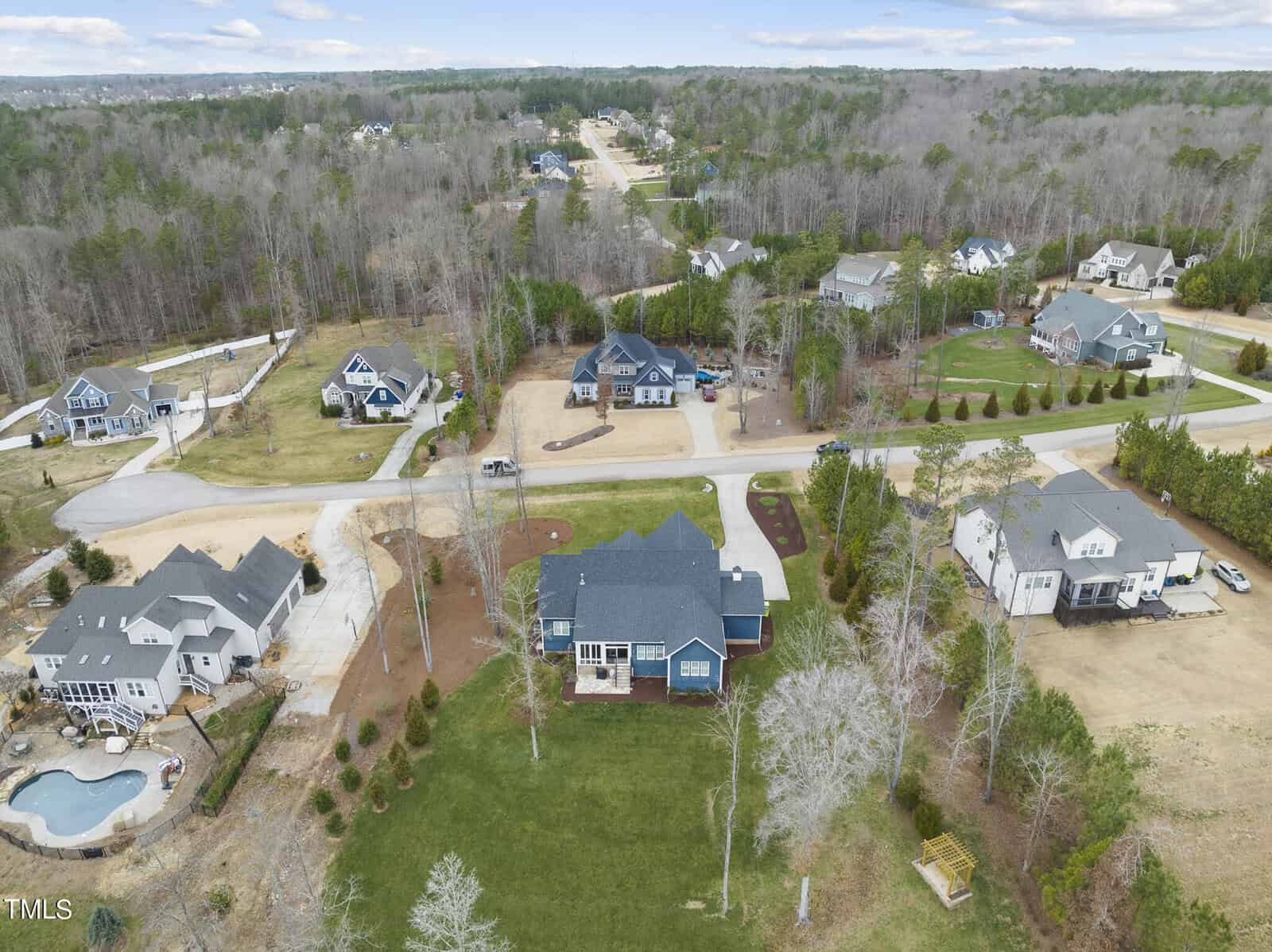 Aerial view of a suburban neighborhood featuring several large, detached houses with spacious yards. The houses are surrounded by trees and grassy areas, with winding streets connecting them. A circular driveway and a swimming pool are visible.