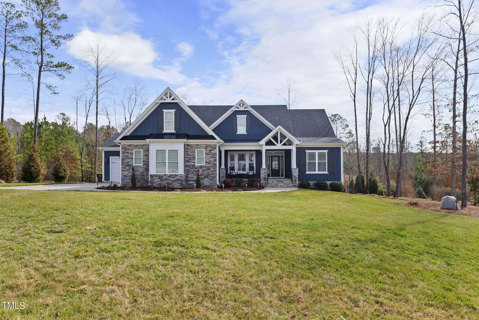 A modern, single-story house with dark blue siding and stone accents, nestled in a grassy area with sparse trees. It has white trim and a large front porch. The sky is partly cloudy.