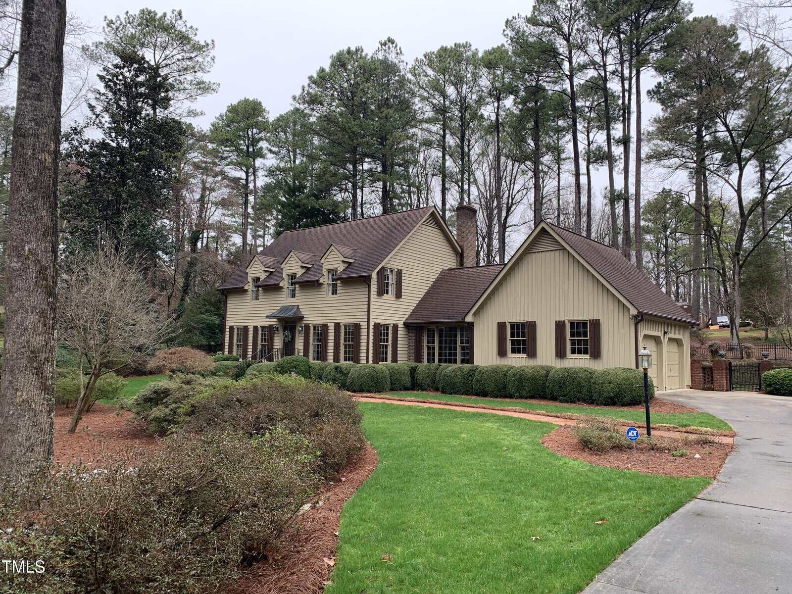 A two-story house at 1750 Parker Lane with beige siding and dark brown shutters surrounded by tall pine trees and manicured landscaping, featuring a curved red pathway and overcast sky.