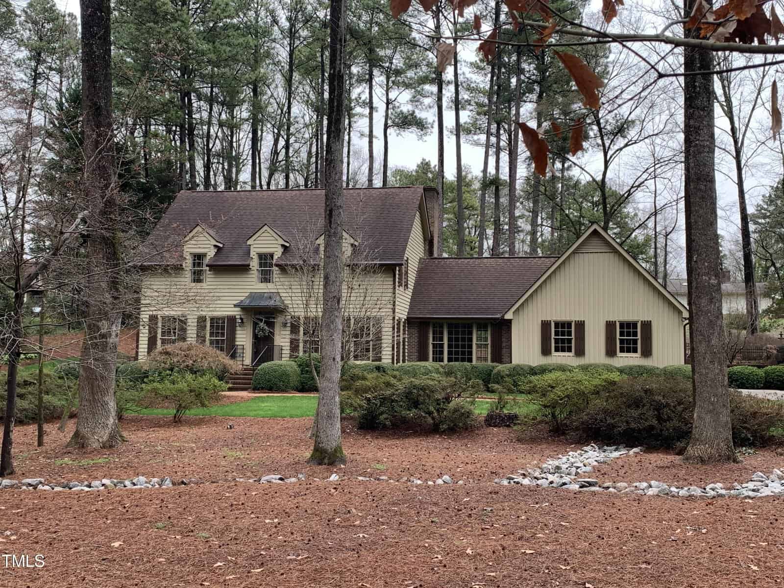 A two-story beige house with a dark roof, surrounded by tall pine trees and a manicured garden with a white pebble path in a suburban setting.