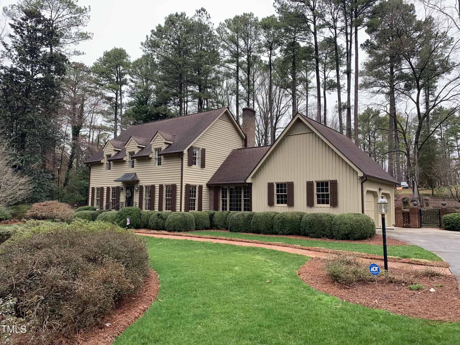 A two-story beige house with dark brown shutters and multiple gabled roofs, surrounded by a manicured lawn and lush greenery, under an overcast sky.