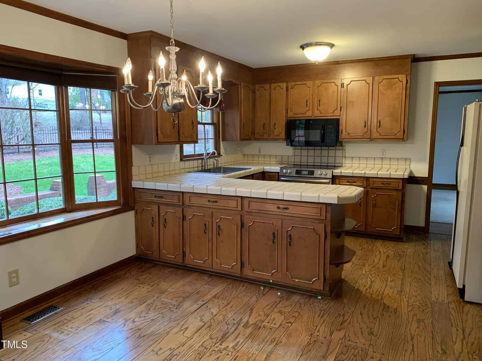 A spacious kitchen with wooden cabinetry, white countertops, and built-in appliances. includes a chandelier, hardwood floors, and a view to a green backyard through a window.