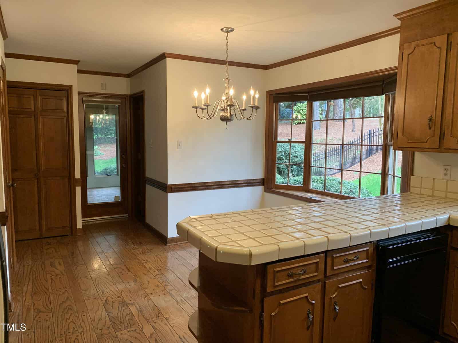 A traditional kitchen featuring wooden cabinetry, white tiled countertops, hardwood flooring, and a chandelier. two brown wooden doors lead to adjacent rooms, visible through a window.