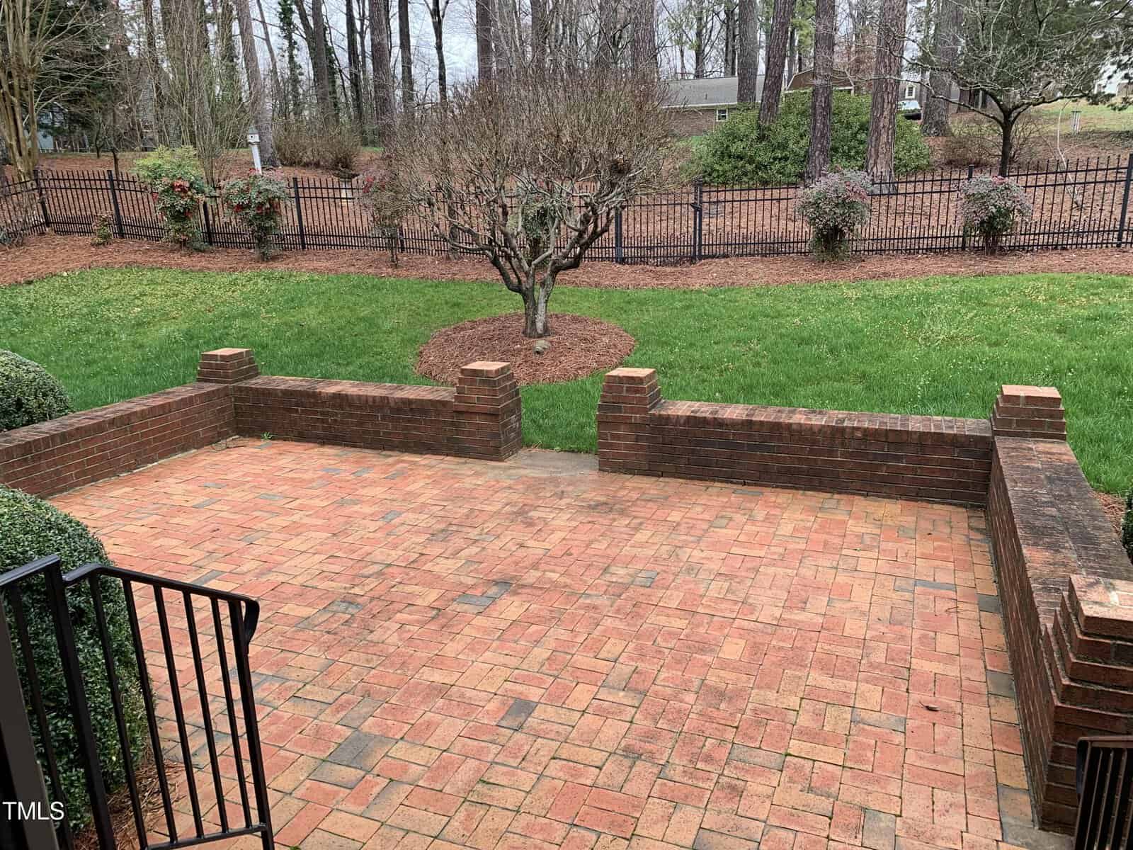 A cozy brick patio surrounded by a low brick wall, overlooking a manicured garden with a central tree and bushes, enclosed by a wrought iron fence under an overcast sky.