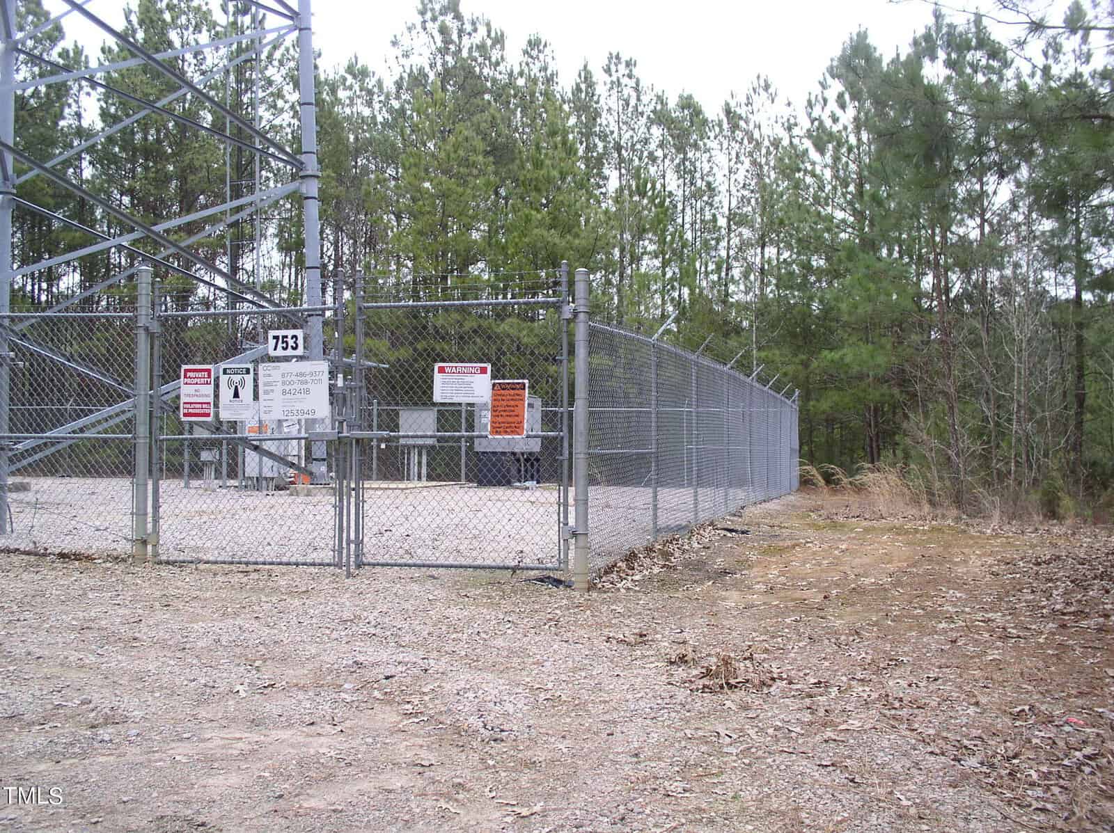 A secured area with a high metal fence and multiple warning signs around the gate, located in a wooded area with a gravel path leading up to it.