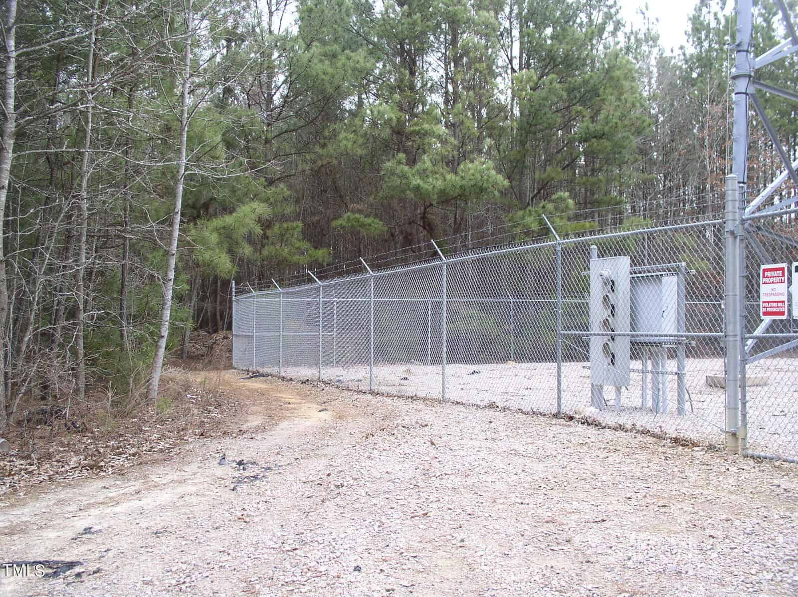 A chain-link fence with a secure gate encloses an area surrounded by pine trees, with a gravel pathway leading along the side. a "no trespassing" sign is visible on the gate.