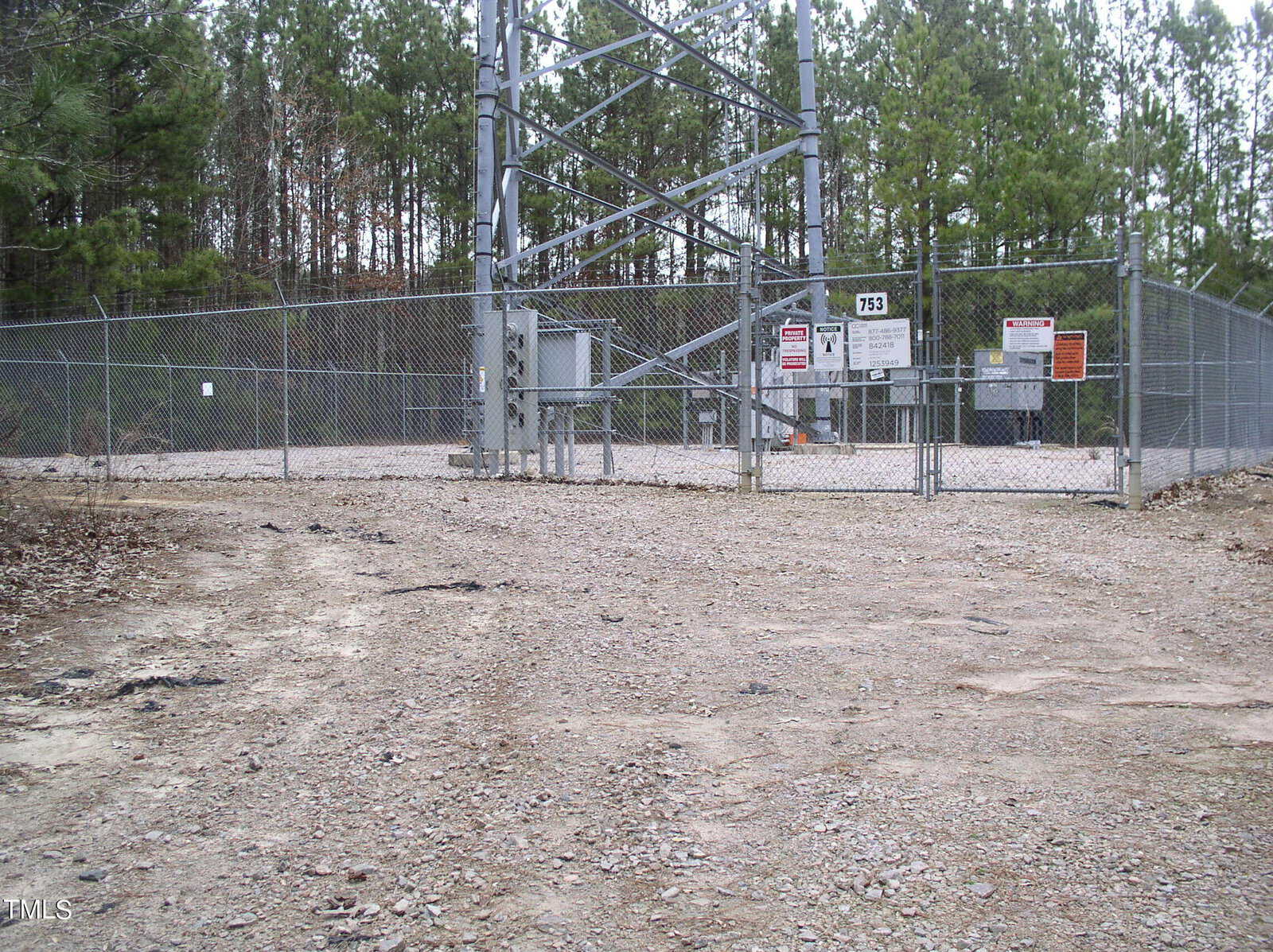 An image showing a gated electrical substation with multiple warning signs, surrounded by a chain-link fence in a barren, dirt-covered area with sparse vegetation.