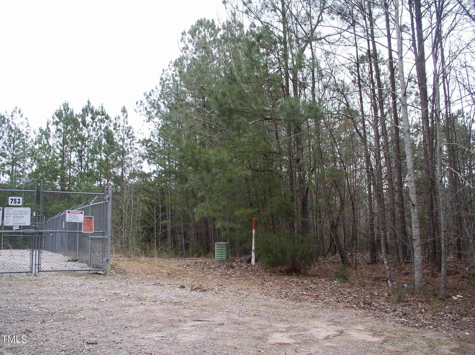 A fenced-off area with a chain-link gate on a barren ground, surrounded by tall pine trees, under a cloudy sky. a sign on the fence displays "for sale" information.