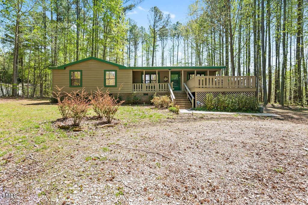 A single-story green house with white trim, featuring a large front porch and a wooden deck, surrounded by tall pine trees under a clear sky. there is a gravel driveway and sparse vegetation in the front yard.