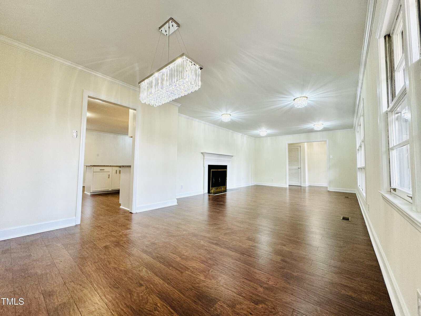 An empty, spacious room with glossy hardwood floors, white walls, and multiple windows allowing abundant natural light. features include a chandelier, recessed lighting, and a fireplace in the background.
