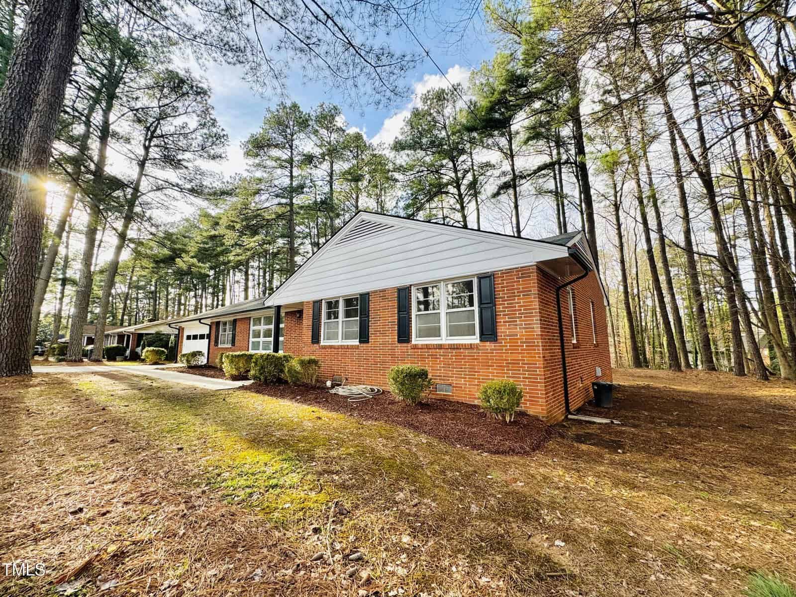 A single-story brick house with a gray roof, located in a sunny wooded area with tall pine trees, featuring a spacious yard and clear blue sky.