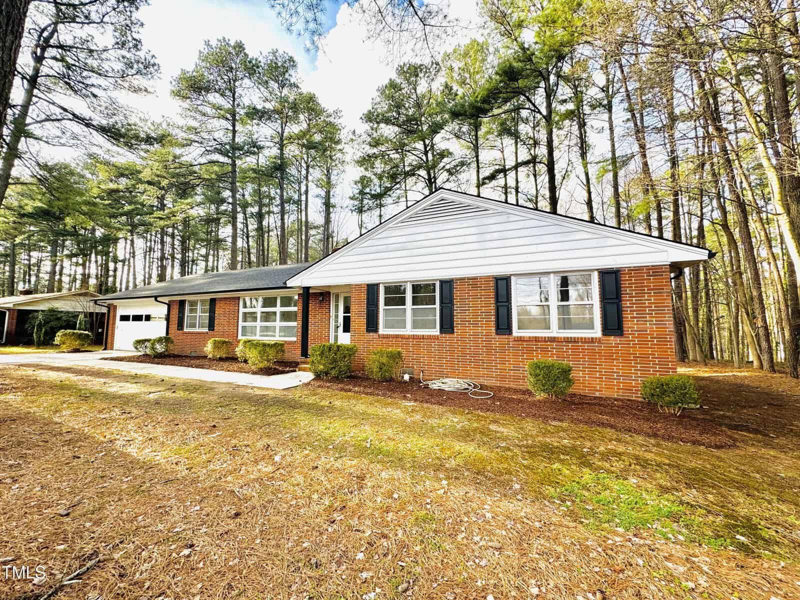 A single-story brick house with a white roof and attached carport, surrounded by a wooded area with tall pine trees under a partly cloudy sky.