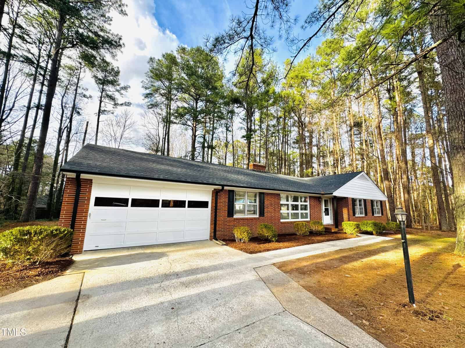 A single-story brick house at 2330 Oxford Road, Henderson NC, with a white double garage and sloping roof, surrounded by tall pine trees under a clear sky. A concrete driveway leads to