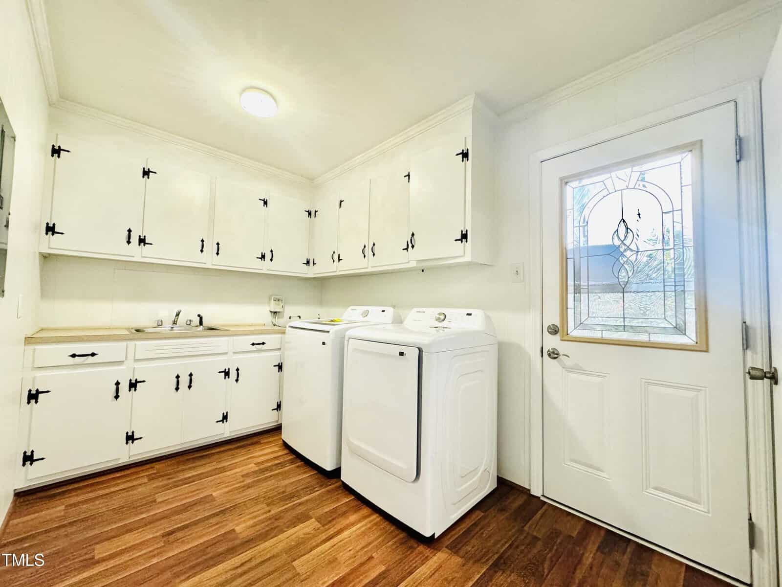 A bright, clean laundry room with white cabinetry and appliances, featuring a decorative glass door and wooden flooring.