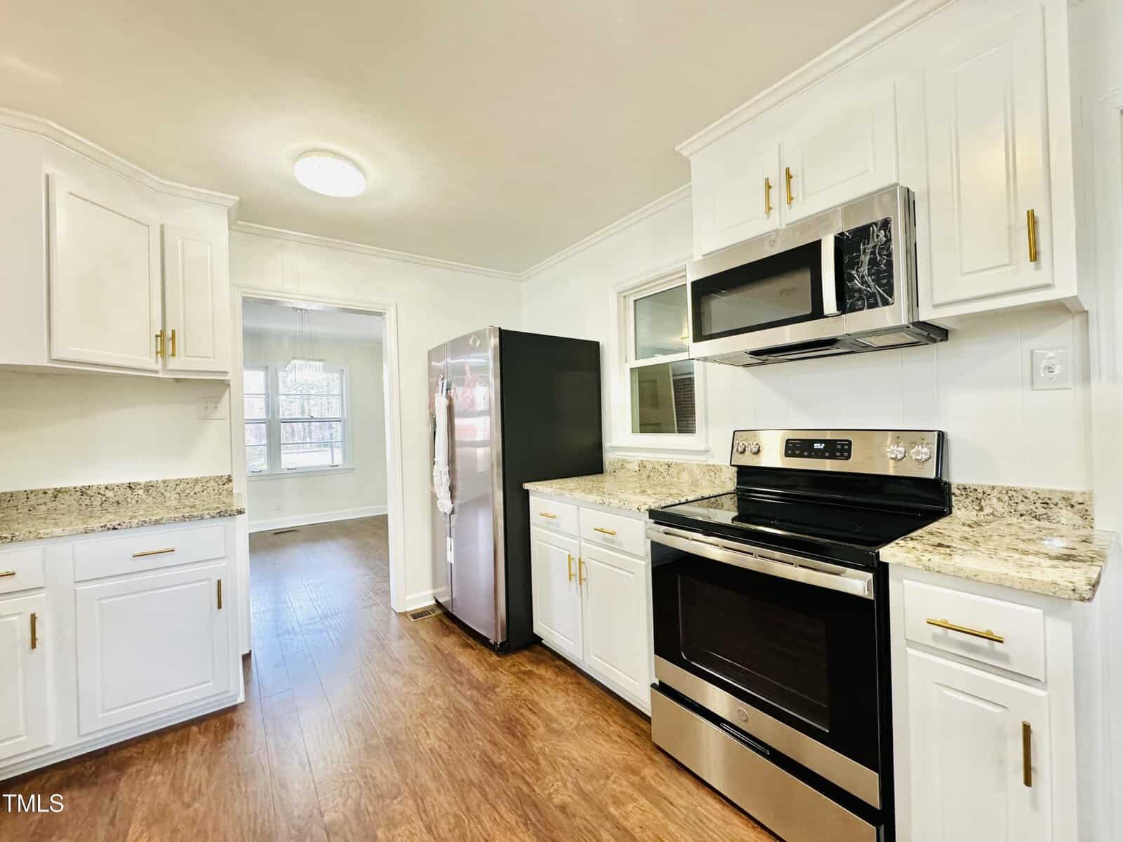A bright, modern kitchen with white cabinetry, granite countertops, and stainless steel appliances, including a refrigerator and oven. there's a window above the sink and hardwood flooring.