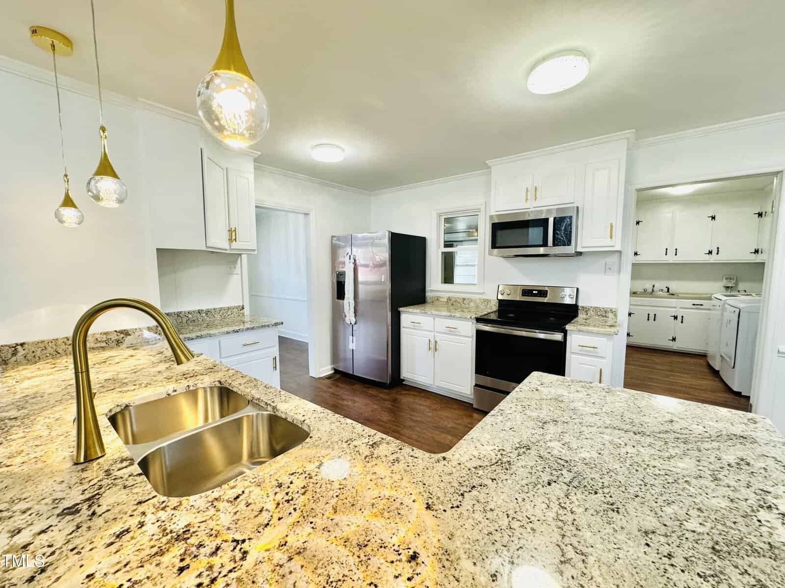 A modern kitchen with white cabinetry, granite countertops, and stainless steel appliances including a fridge, microwave, and oven. two pendant lights hang above.