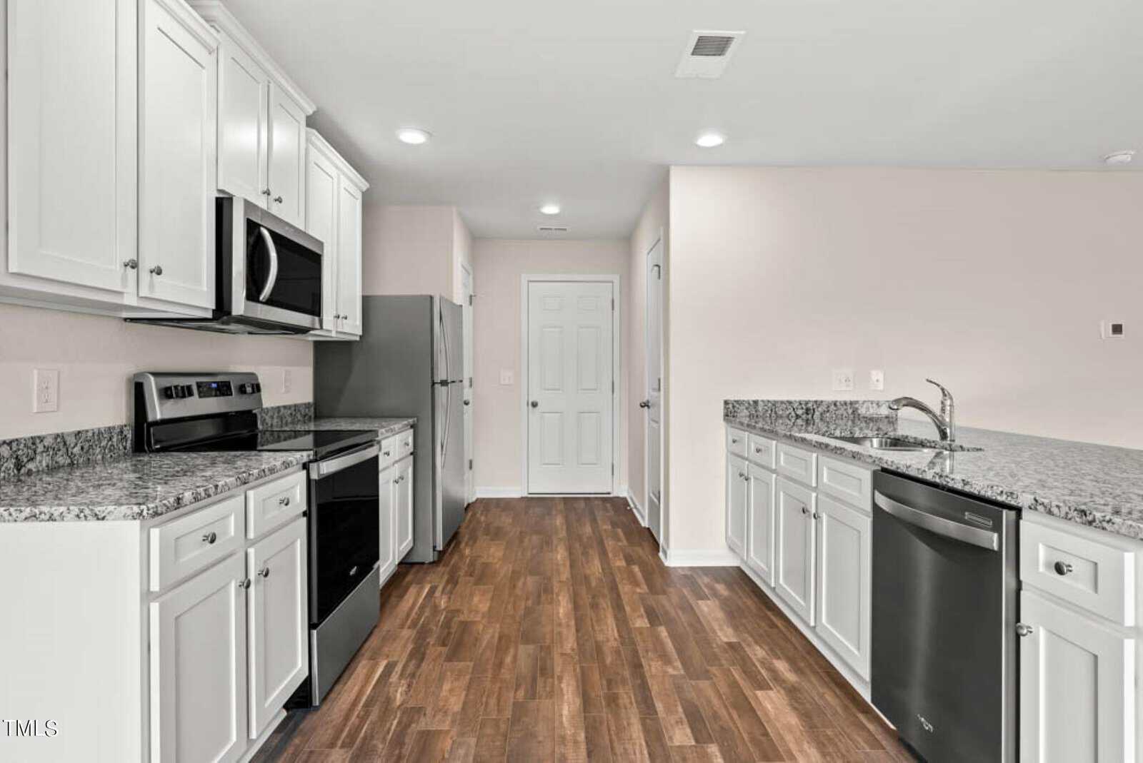 A modern kitchen with white cabinets, stainless steel appliances, and gray granite countertops. There's a double sink, stove, microwave, refrigerator, and dishwasher. The floor is a dark wood laminate. A door is at the far end of the room.