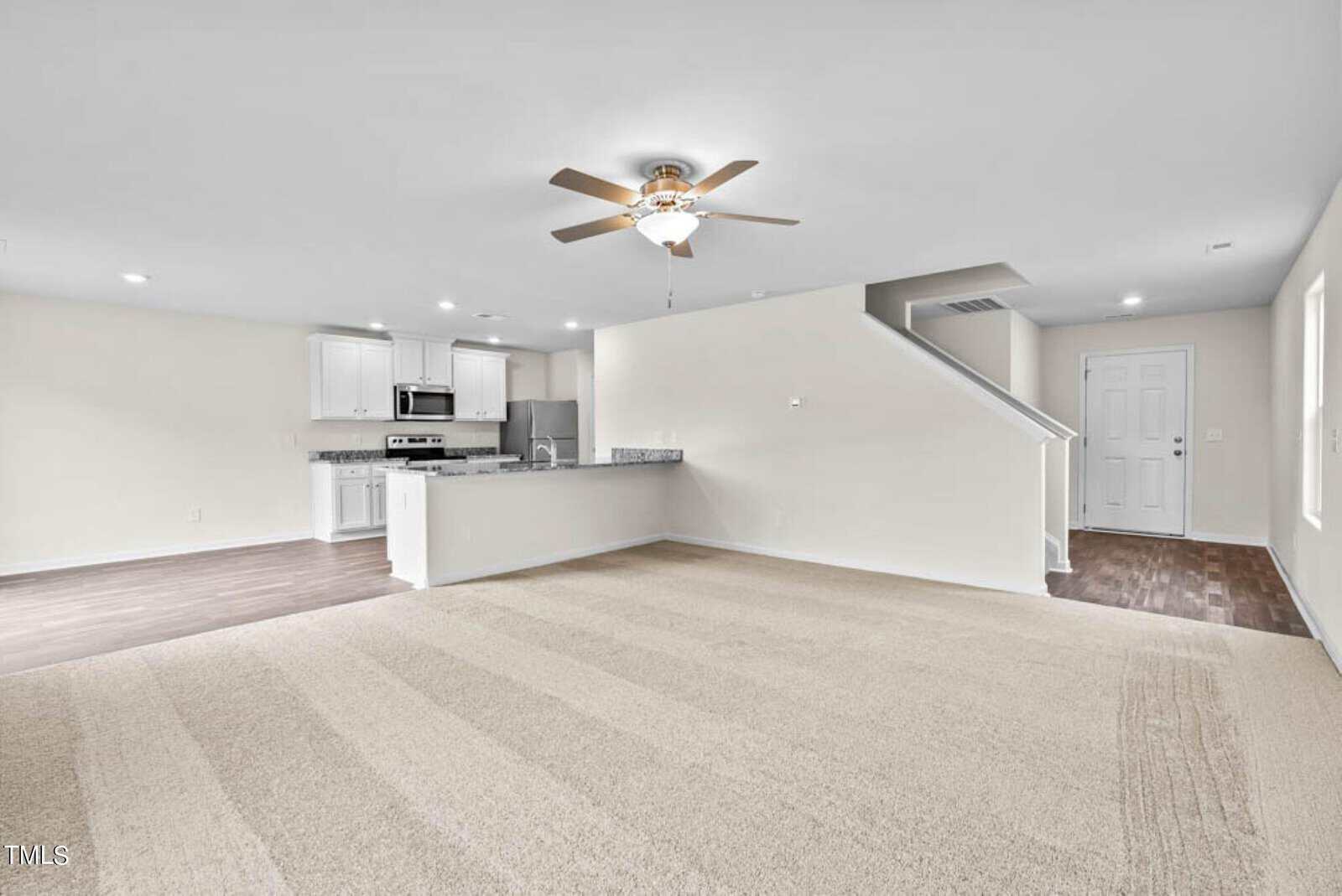 Interior of a modern, unfurnished open-concept living room with beige carpet. Includes a ceiling fan, adjacent kitchen with stainless steel appliances, white cabinets, and a staircase leading to the second floor. Brown wood flooring in kitchen area.