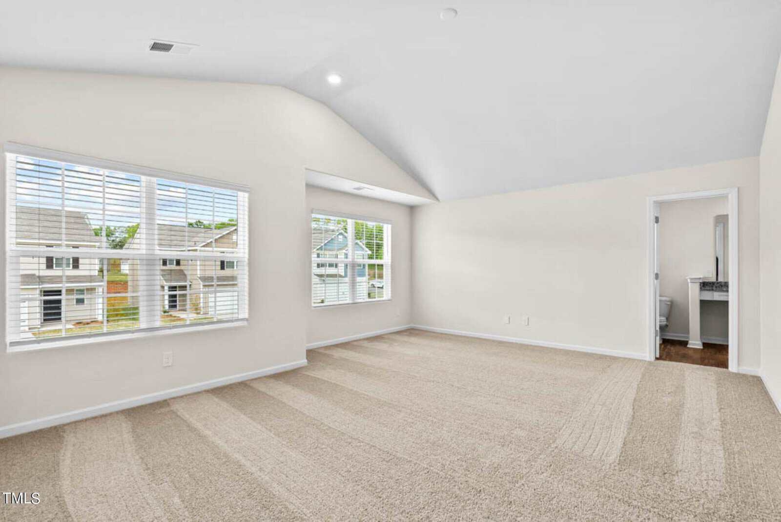 Spacious, empty, carpeted room with vaulted ceiling, large windows, and an open doorway leading to a bathroom. Natural light fills the space, revealing a view of neighboring houses through the windows. Walls are painted light cream.