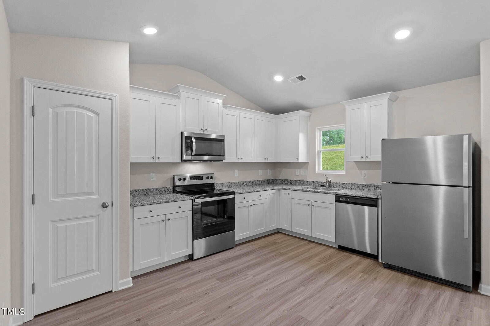 A modern kitchen with white cabinets, stainless steel appliances, and granite countertops. The room has light wood flooring, a window above the sink, and recessed lighting in the ceiling.