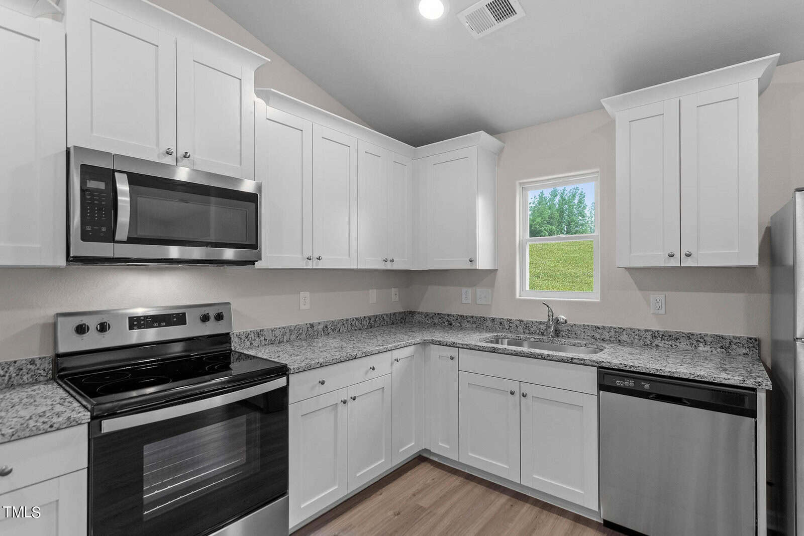 A modern kitchen with white cabinets, granite countertops, and stainless steel appliances, including a microwave, oven, and dishwasher. A small window lets in natural light, and the floor is wooden.