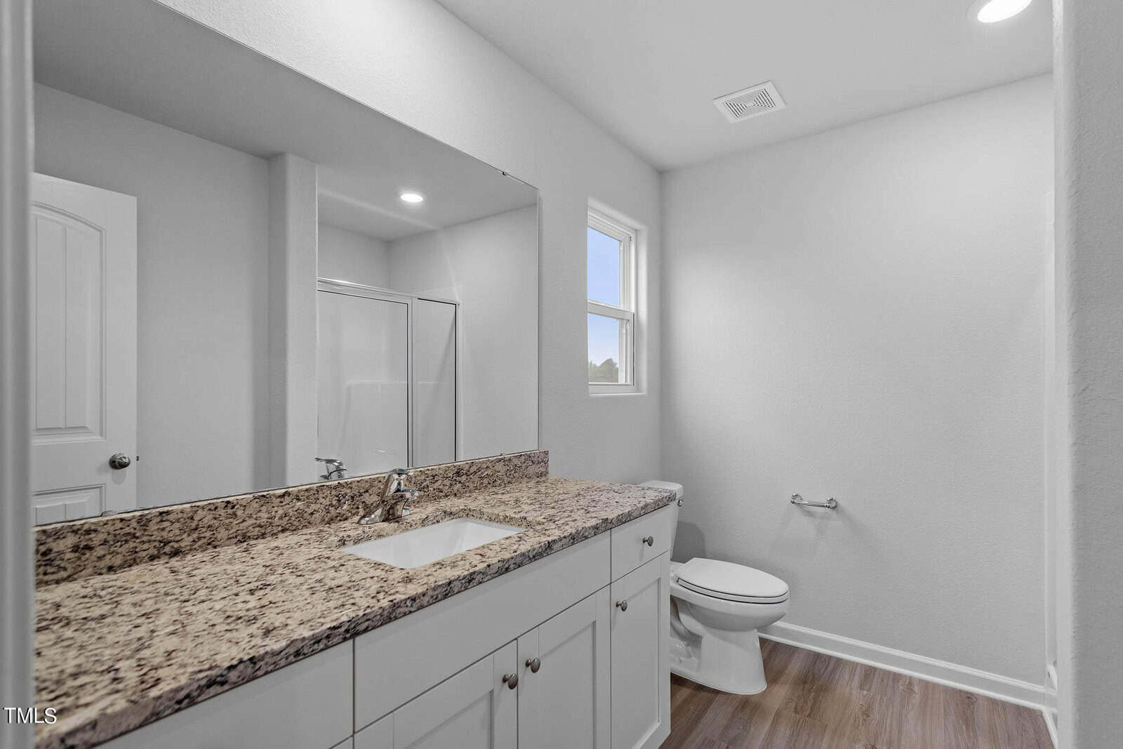 A modern bathroom with a granite countertop and white cabinets under a large mirror. There's a toilet beside a small window, and a shower stall with a glass door, all set on wood-look flooring. The room is brightly lit.