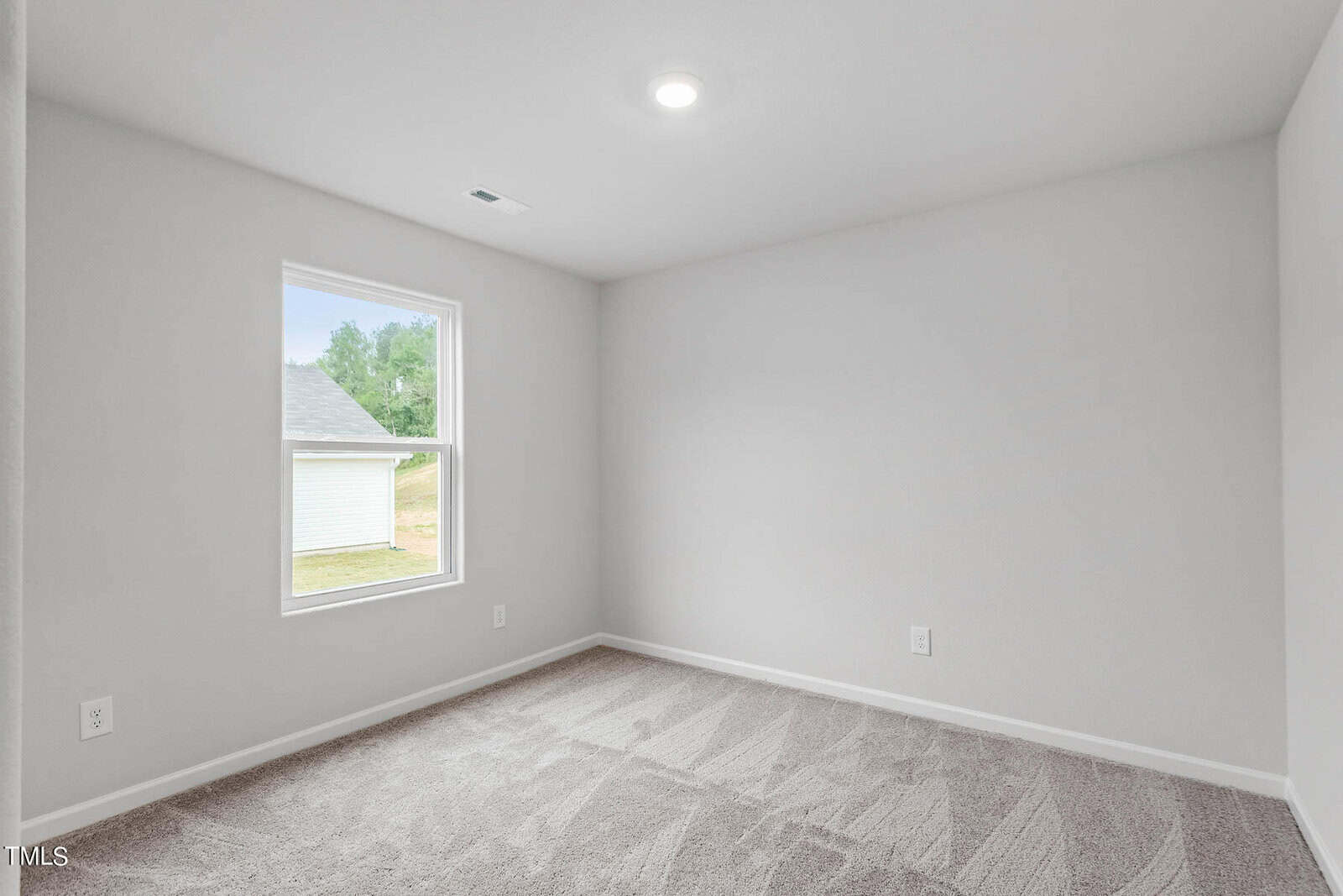 A small, empty room with light gray walls and carpeted floor. There is a single window on the left side, allowing natural light in. The ceiling features a recessed light. The walls and room appear freshly painted and clean.