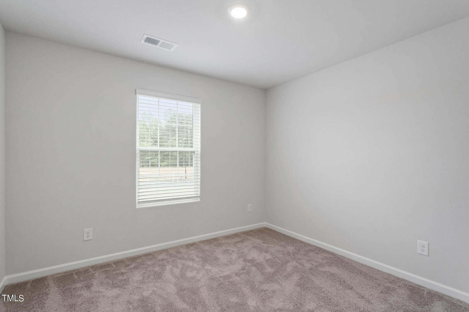 A small, empty room with light gray walls and a beige carpet. A single window on the left wall lets in natural light. The ceiling has a recessed light, and there's a square air vent above the window.