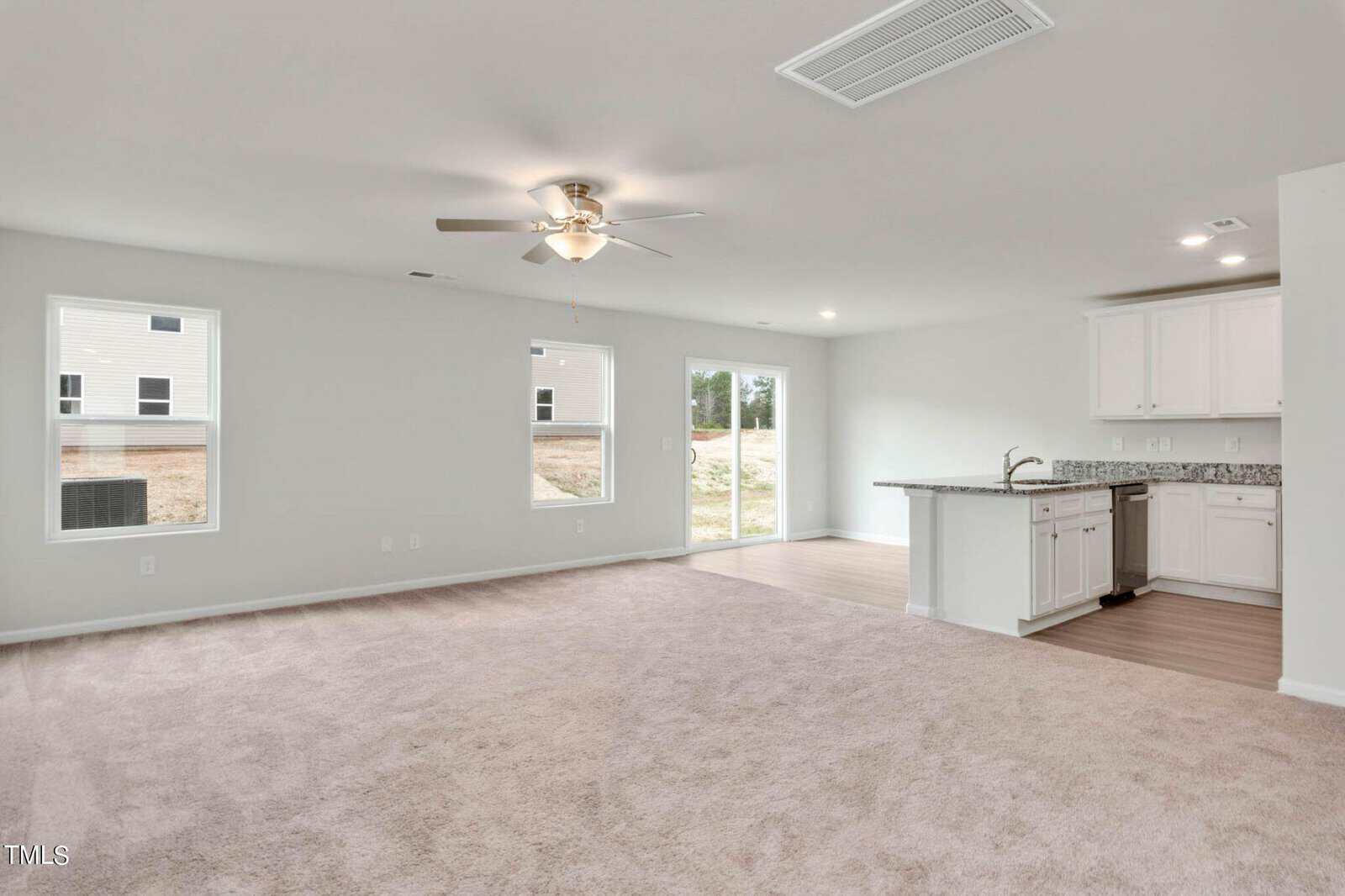 An empty living room with beige carpet and a ceiling fan. Adjacent is a kitchen area with white cabinets, a granite countertop, and a sink. Large windows and a sliding glass door provide natural light from outside.