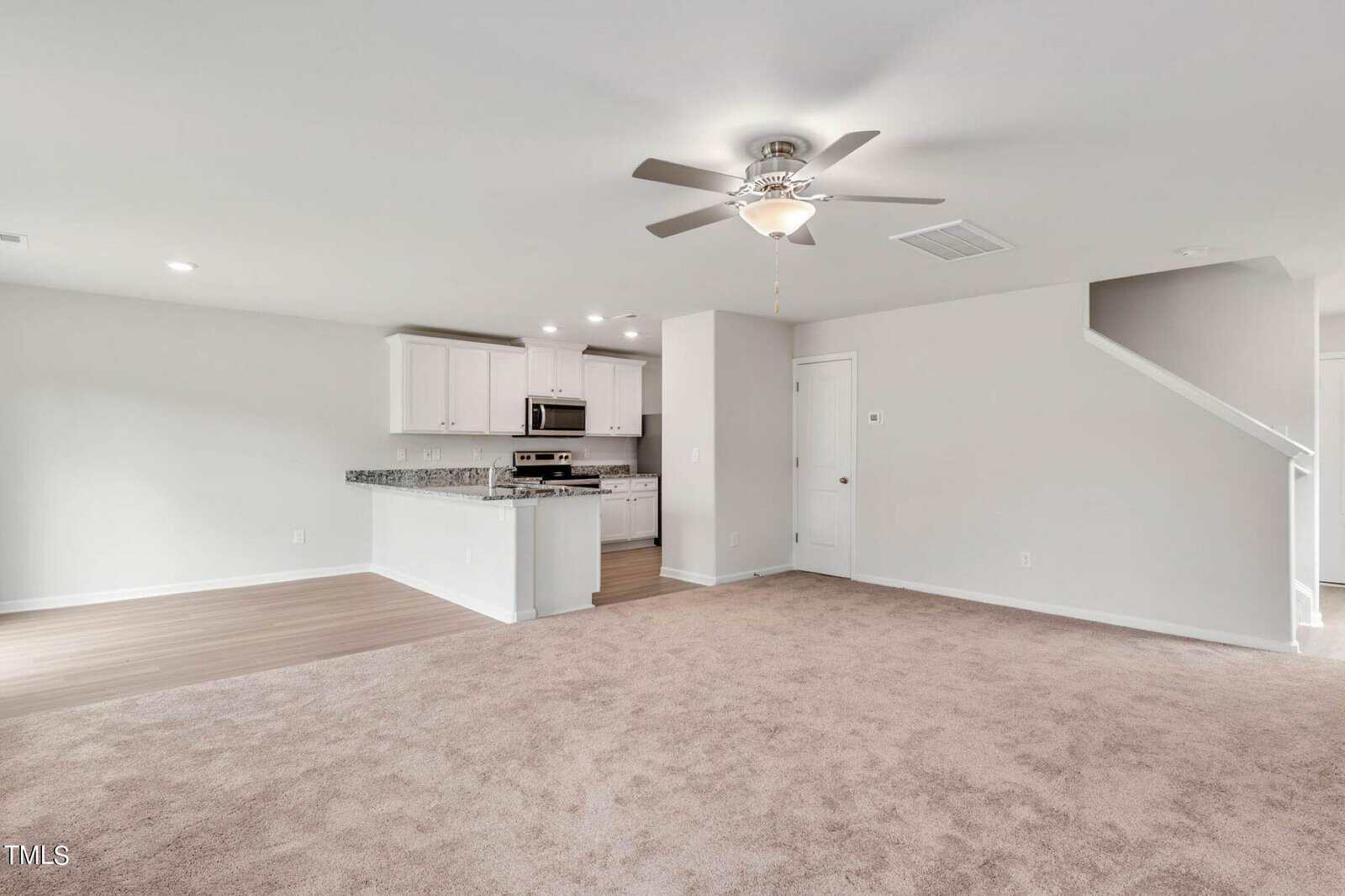 Spacious open-plan living area with beige carpet and light gray walls. Modern kitchen with white cabinets, granite countertops, and stainless steel appliances. A ceiling fan is mounted overhead, and a staircase leads to the upper floor.