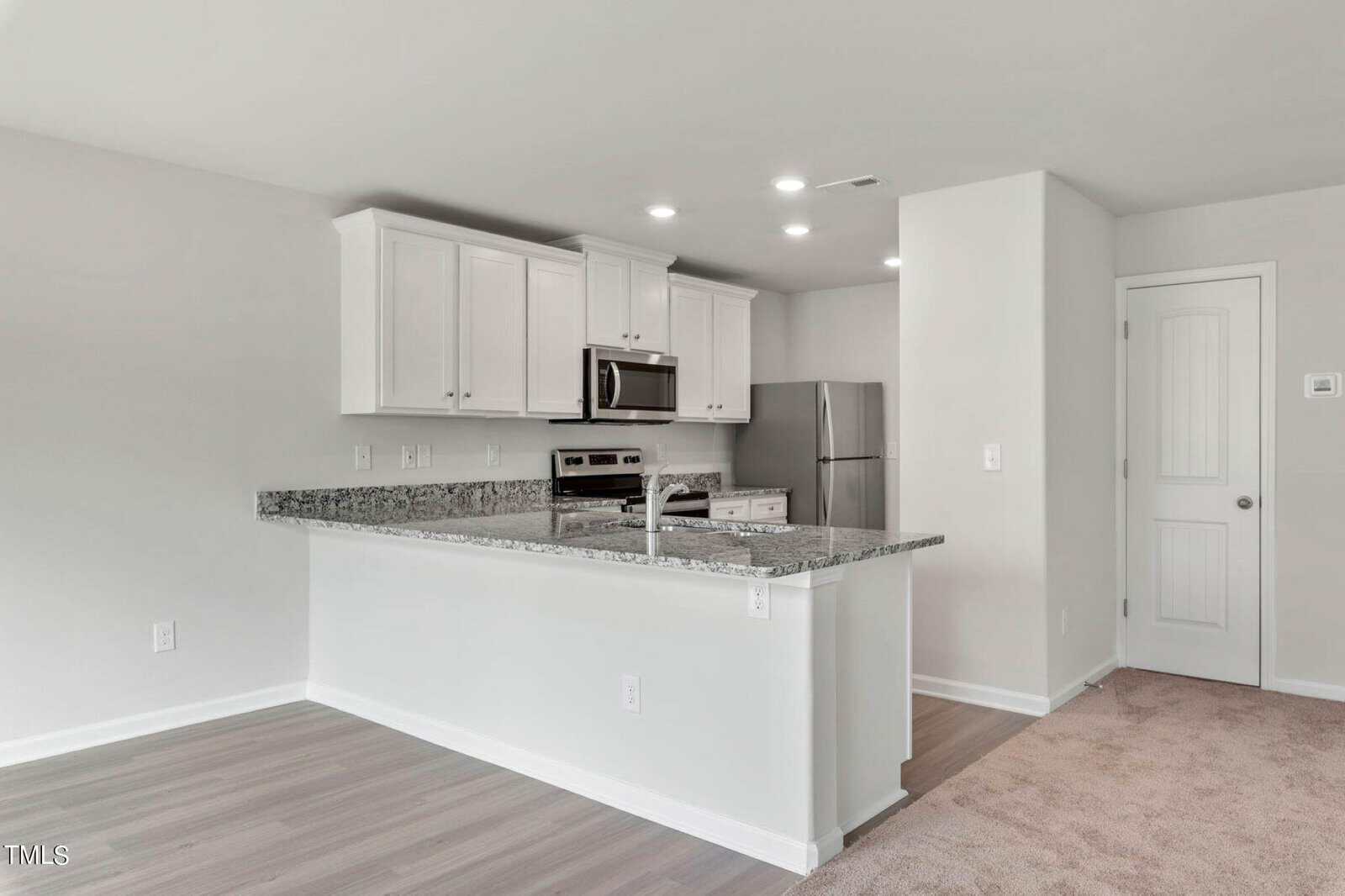 A modern kitchen with white cabinets, stainless steel appliances, and granite countertops. The kitchen features a bar-height counter separating it from a gray-toned laminated flooring area, adjacent to a carpeted space. Walls are painted light gray.
