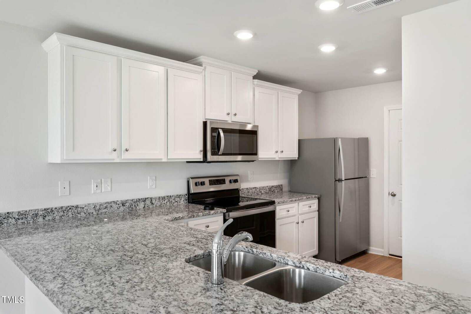 A modern kitchen featuring white cabinets, a stainless steel refrigerator, a built-in microwave, and an electric stove. The foreground shows a granite countertop with a dual sink, and recessed lighting illuminates the space.