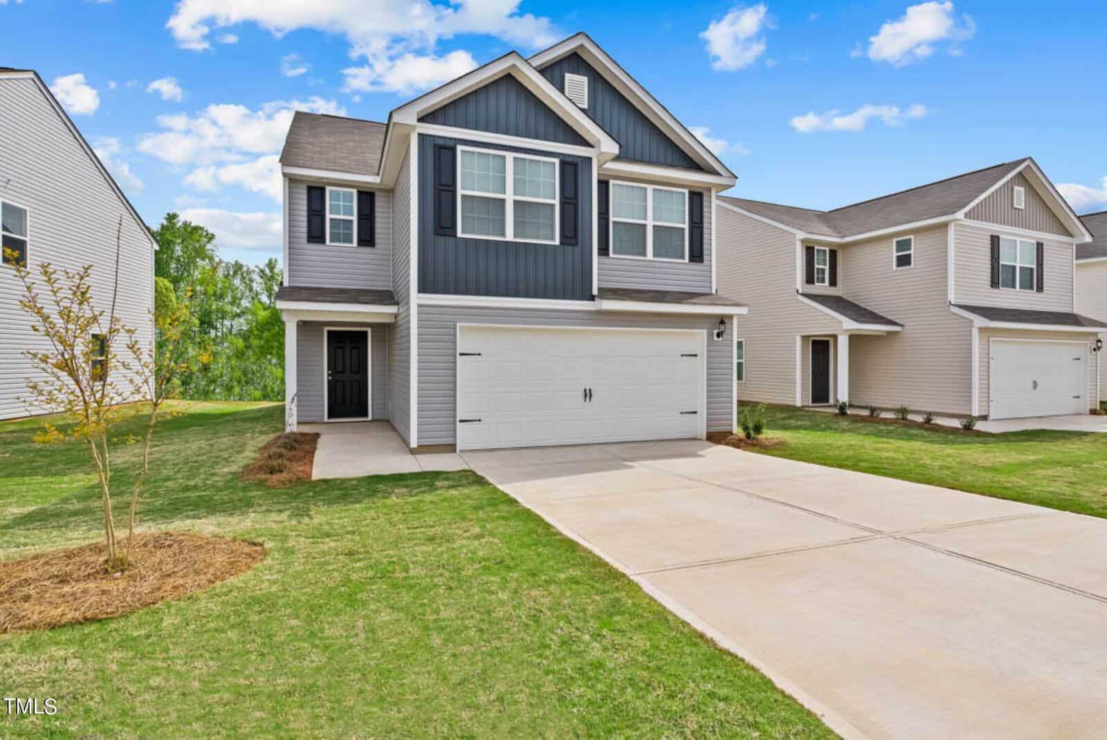 A two-story suburban house on 100 Scoville Street in Oxford boasts gray siding and white trim, complemented by a double garage and a neatly maintained lawn. The sky is blue with scattered clouds, and nearby houses are visible.