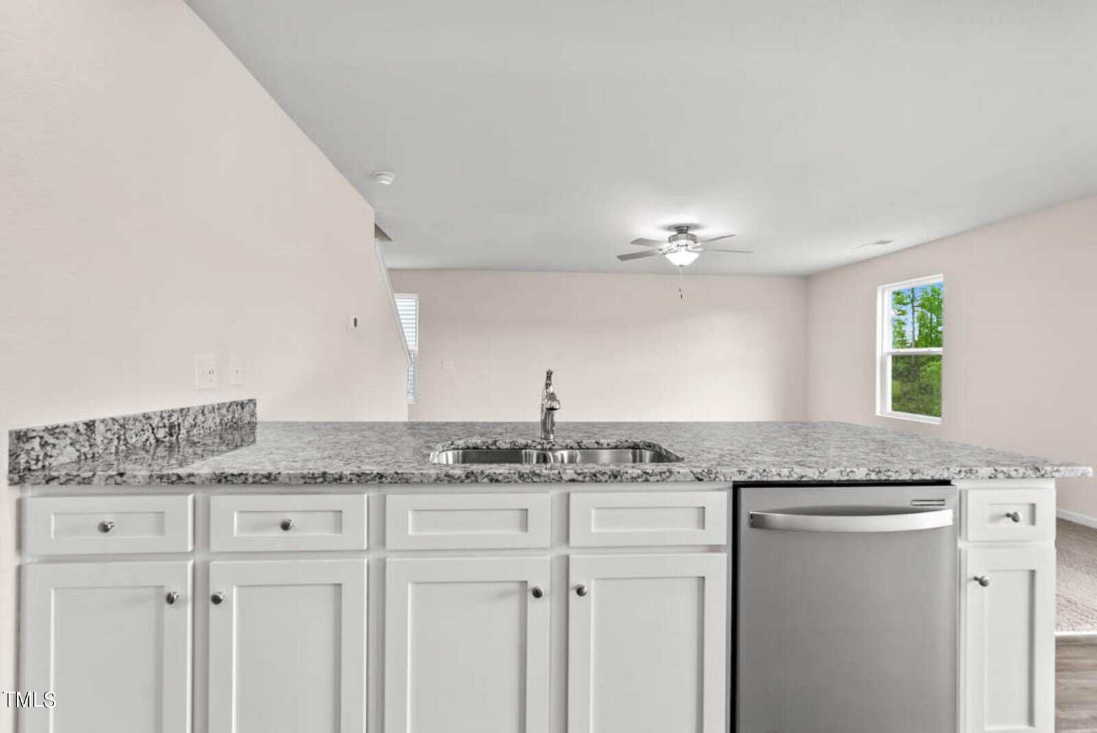 Modern kitchen interior with a large granite countertop island featuring a sink and stainless steel dishwasher. White cabinets line the island. The open-plan space has light walls, and a glimpse of a ceiling fan in the adjacent room is visible.