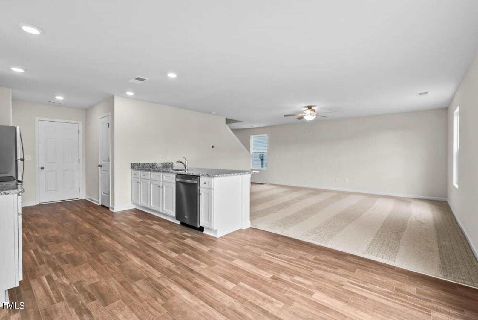 Spacious, open-concept room with wood and carpet flooring. Features a modern kitchen with white cabinetry, granite countertops, and stainless steel appliances. A ceiling fan is visible in the carpeted living area. Bright, neutral walls and ample lighting.