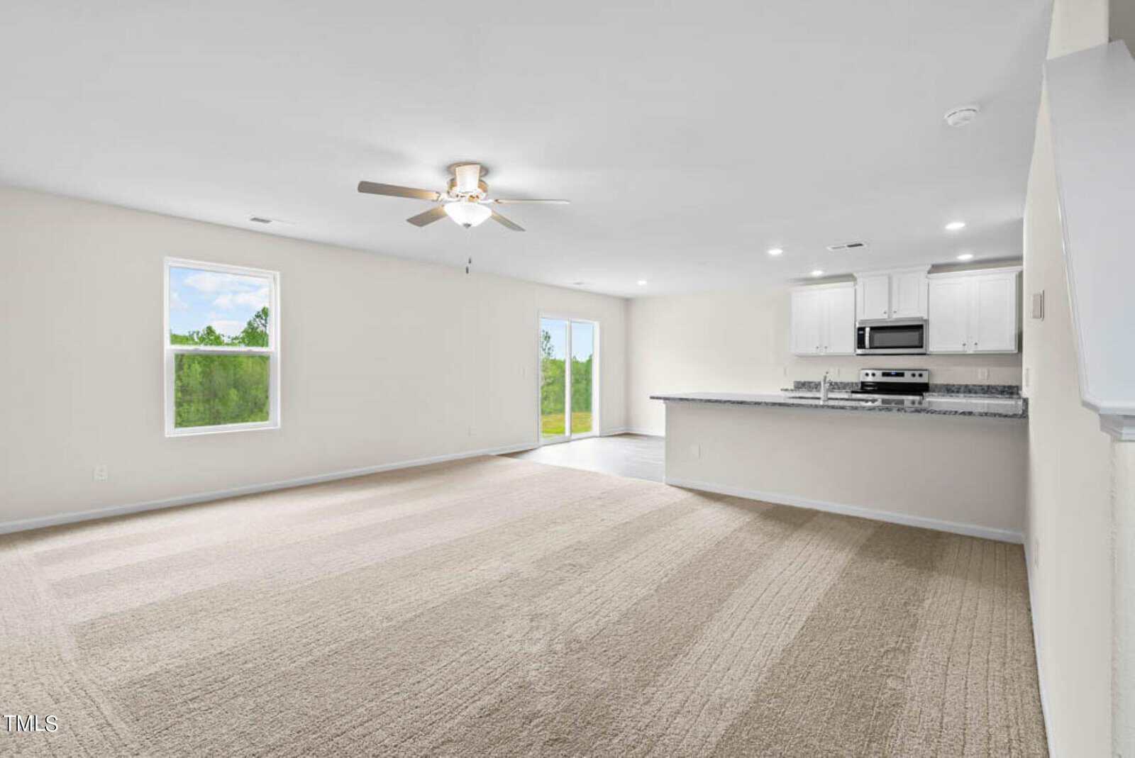 Spacious open-concept living area with beige carpet, light walls, ceiling fan, and recessed lighting. Features a modern kitchen with white cabinets, stainless steel appliances, and a granite countertop with bar seating. Large windows and glass doors offer natural light.