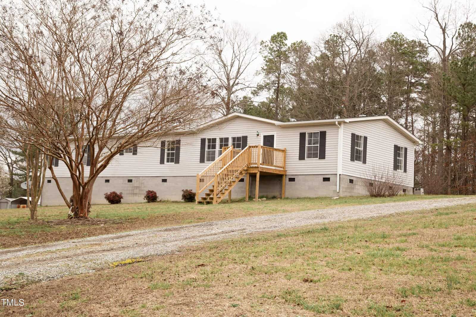 A white modular home with gray roofing, featuring a wooden staircase leading to the front door. it is surrounded by bare trees and a small lawn on a cloudy day.