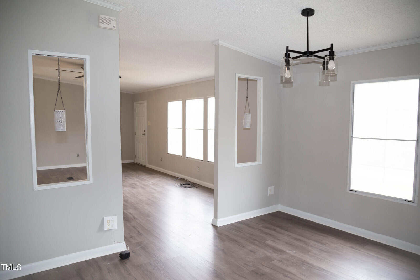 An empty, modern room with gray walls and dark wood flooring. it features a white door, two pendant lights hanging by the hallway, and large windows providing natural light.