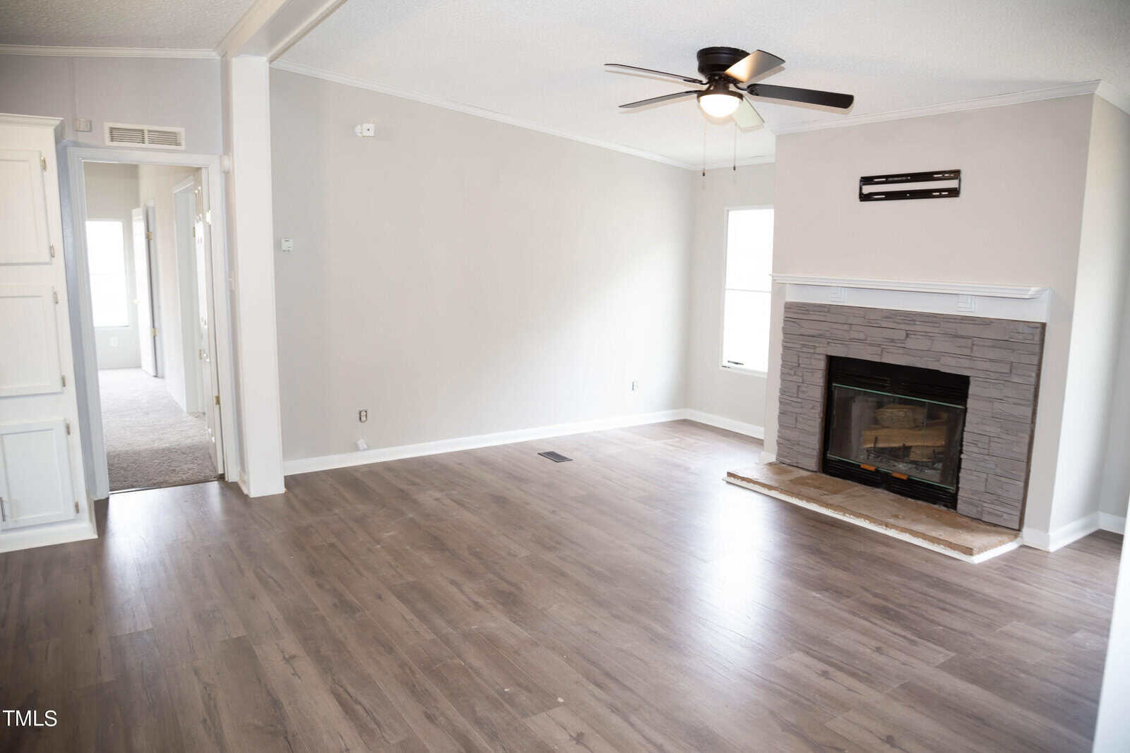 An empty living room with neutral walls, a fireplace surrounded by gray stone, and dark wood floors. the room includes a ceiling fan, a white door on one side, and an air conditioning unit above the fireplace.