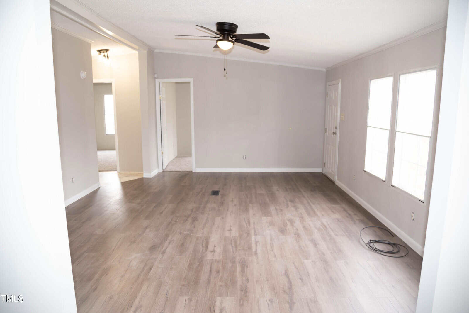 Empty room with gray walls and wooden flooring, featuring a ceiling fan and natural light coming from windows. a door on the far side leads to another room.