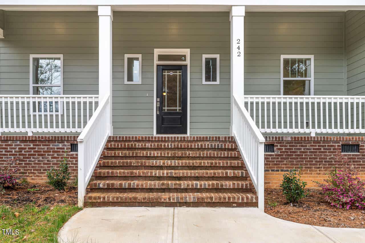 A front porch with brick steps leading to a black door, white railings, and gray siding. The house number 242 is on a column, and small bushes are planted along the brick foundation.
