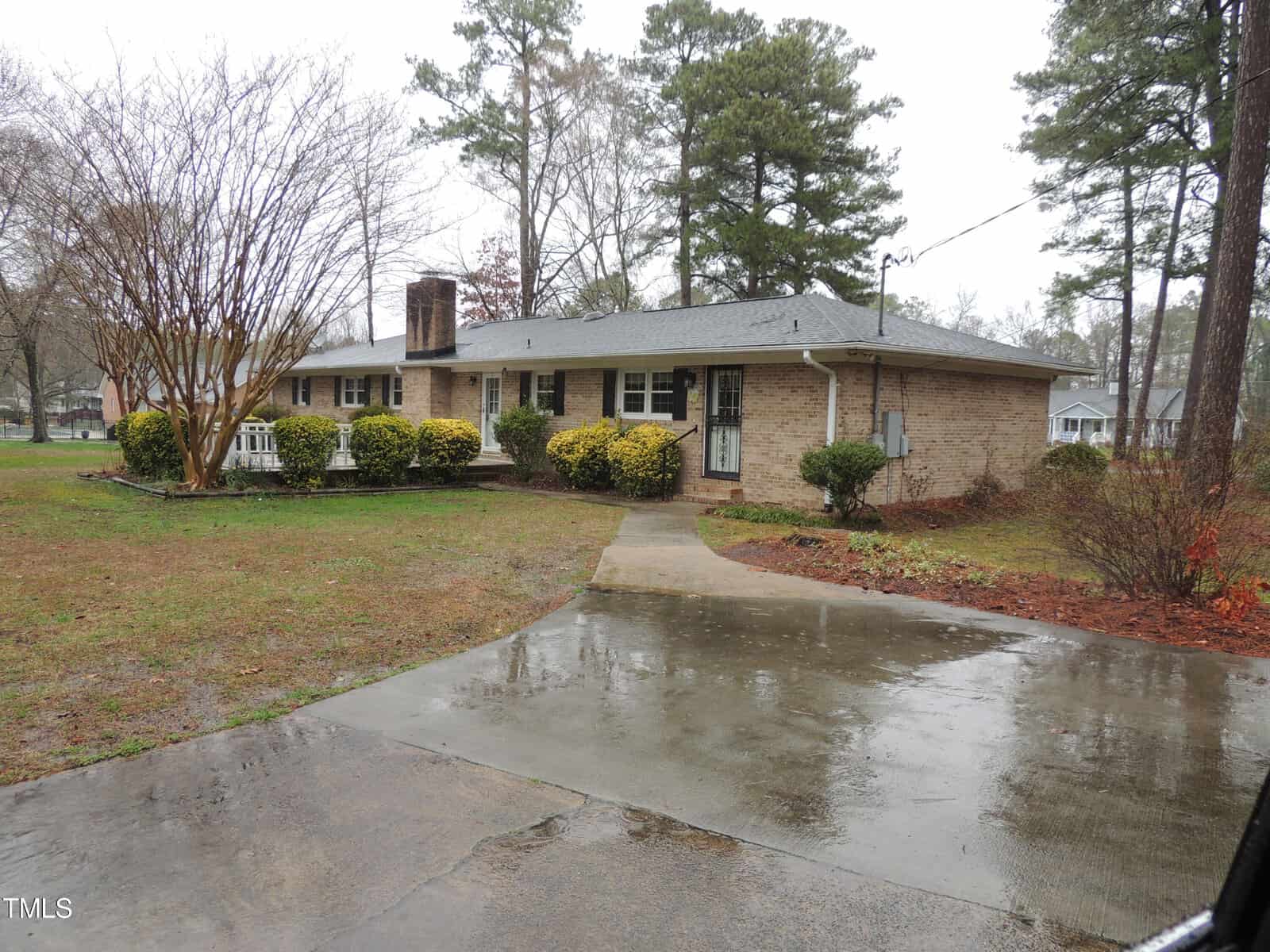 A single-story brick house with a gray roof is surrounded by wet ground, trees, and shrubs. A concrete driveway curves towards the front entrance. The sky appears overcast, suggesting recent rain.