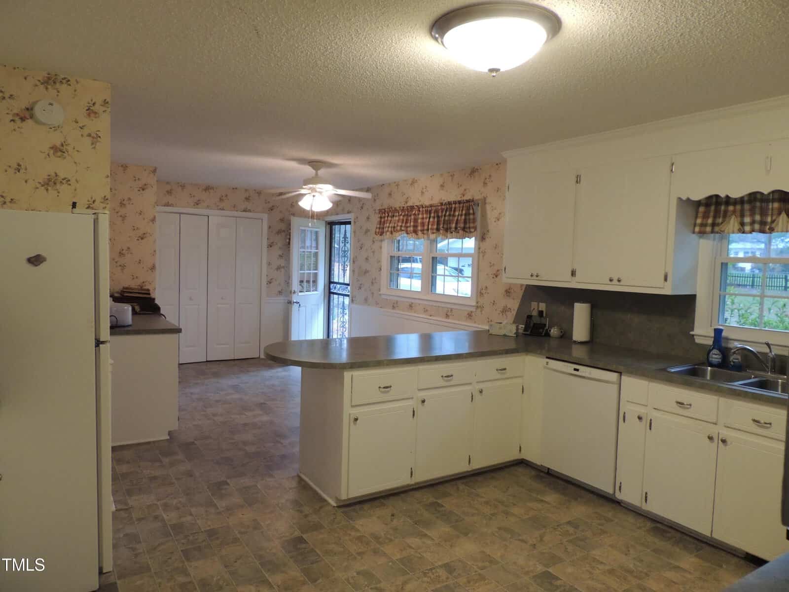 A spacious kitchen with white cabinets and a large island. There's a ceiling fan and large windows with checkered valances. The floor is tiled, and the walls are adorned with floral wallpaper. A door leads to an adjoining room.
