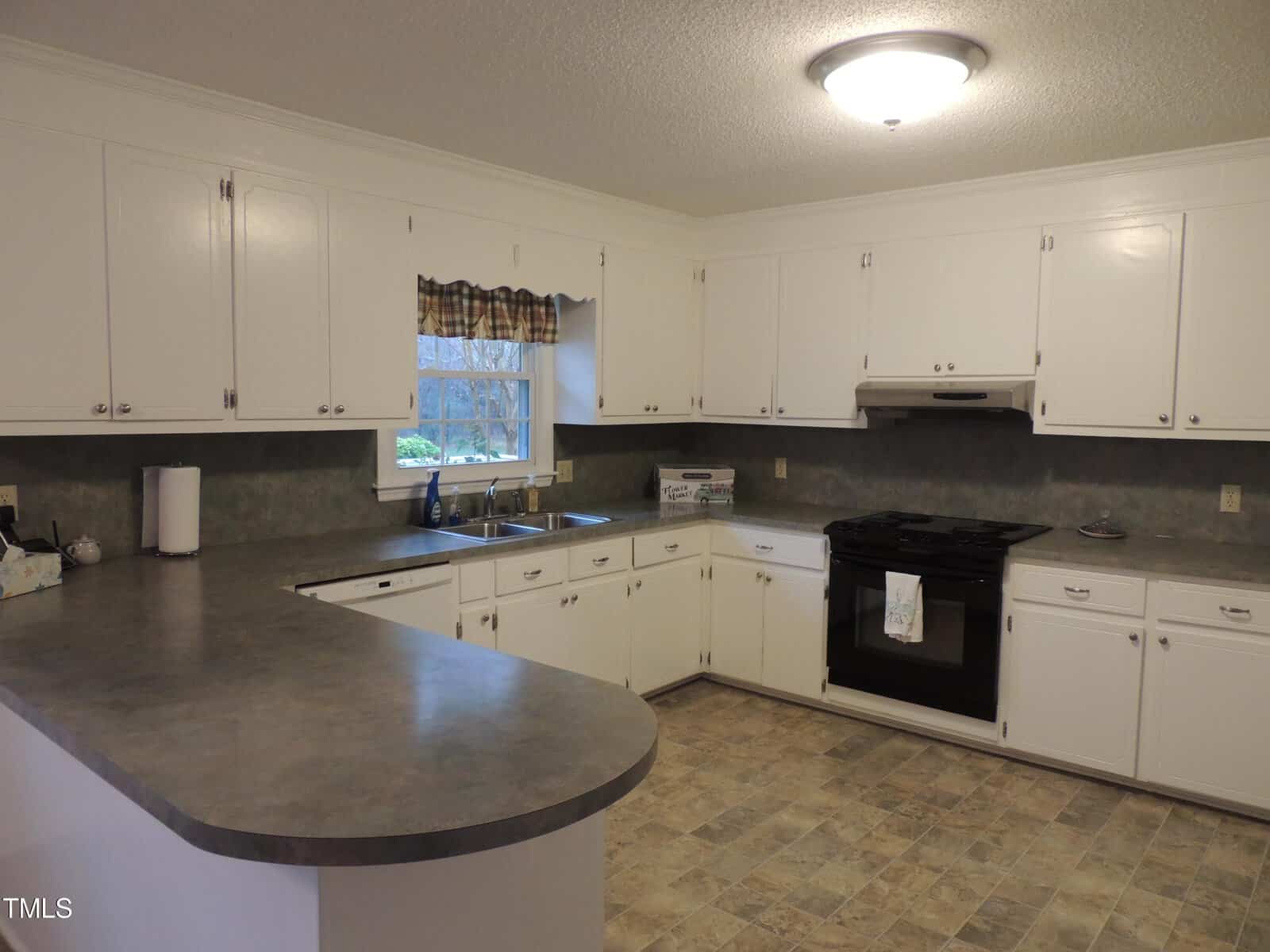 A spacious kitchen with white cabinets, a dark countertop, and a black stove. The room has tiled flooring and a window with a plaid valance above the sink, allowing natural light in. A ceiling light fixture provides additional illumination.
