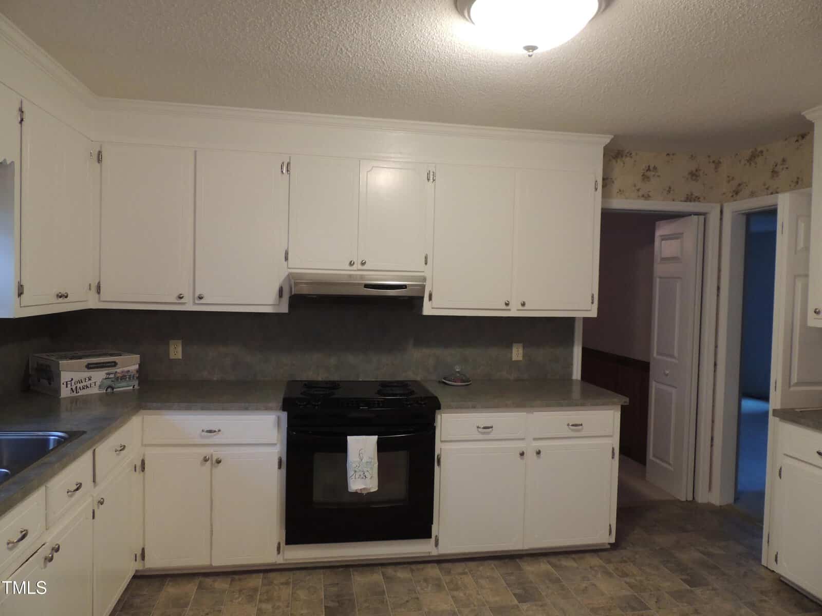 A kitchen with white cabinets, a black electric stove, and gray countertops. A floral wallpaper decorates the walls. The floor is tiled in a neutral tone, and a small window is visible above the sink.