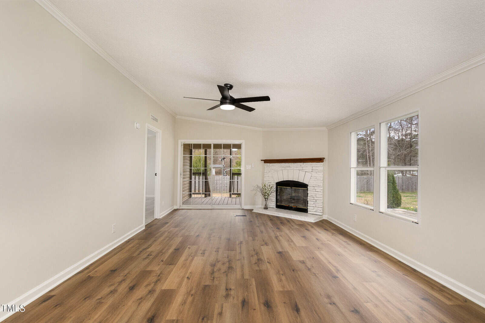 A spacious living room with light wood flooring and cream walls. It features a ceiling fan, a white brick fireplace with a wood beam mantel in the corner, large windows, and a doorway leading to a deck. The room feels bright and airy.