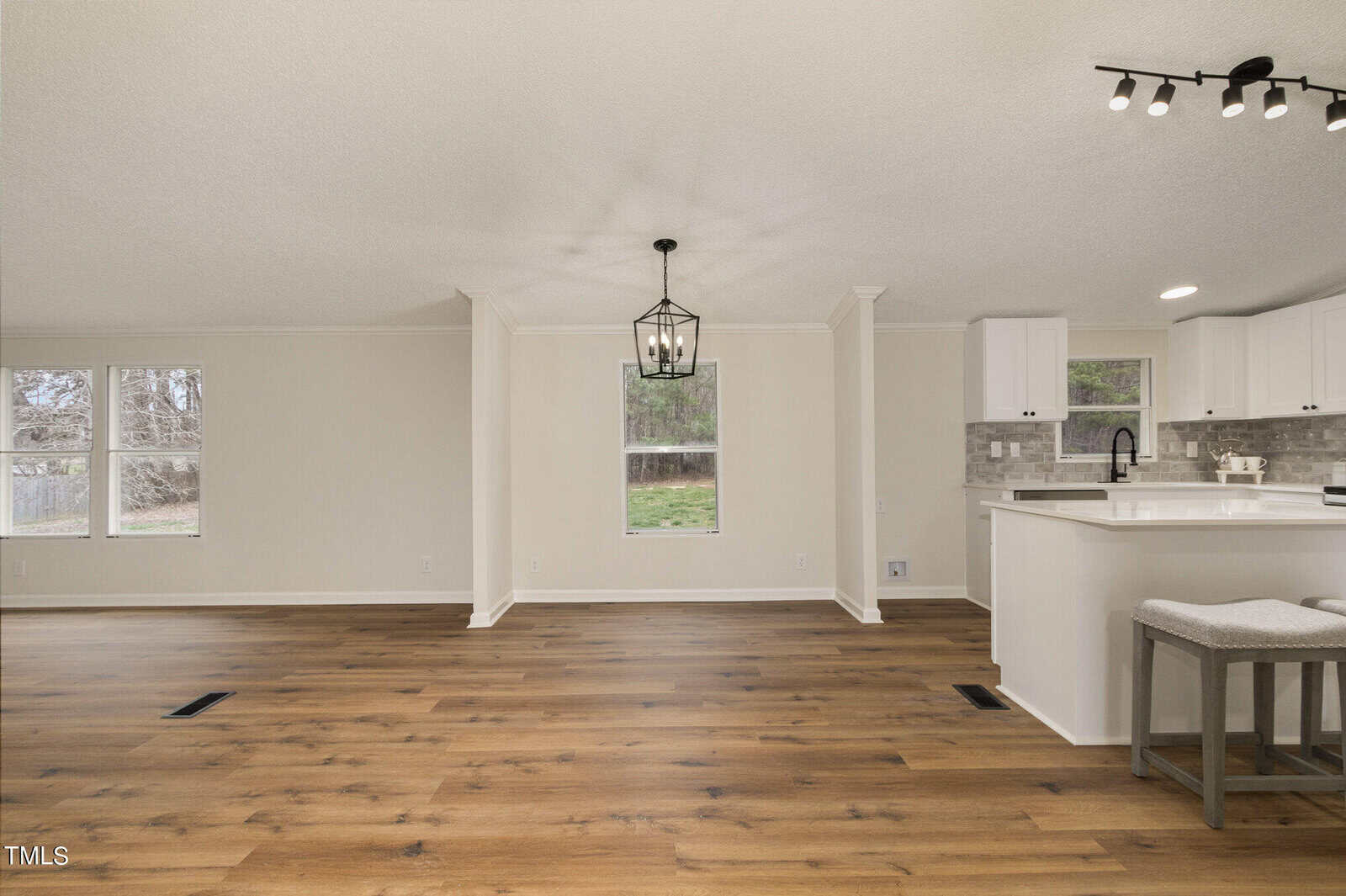 Modern open-concept living area with light wood floors, white walls, and a contemporary black chandelier. The kitchen has white cabinets, a tiled backsplash, and an island with two stools. Large windows provide natural light.