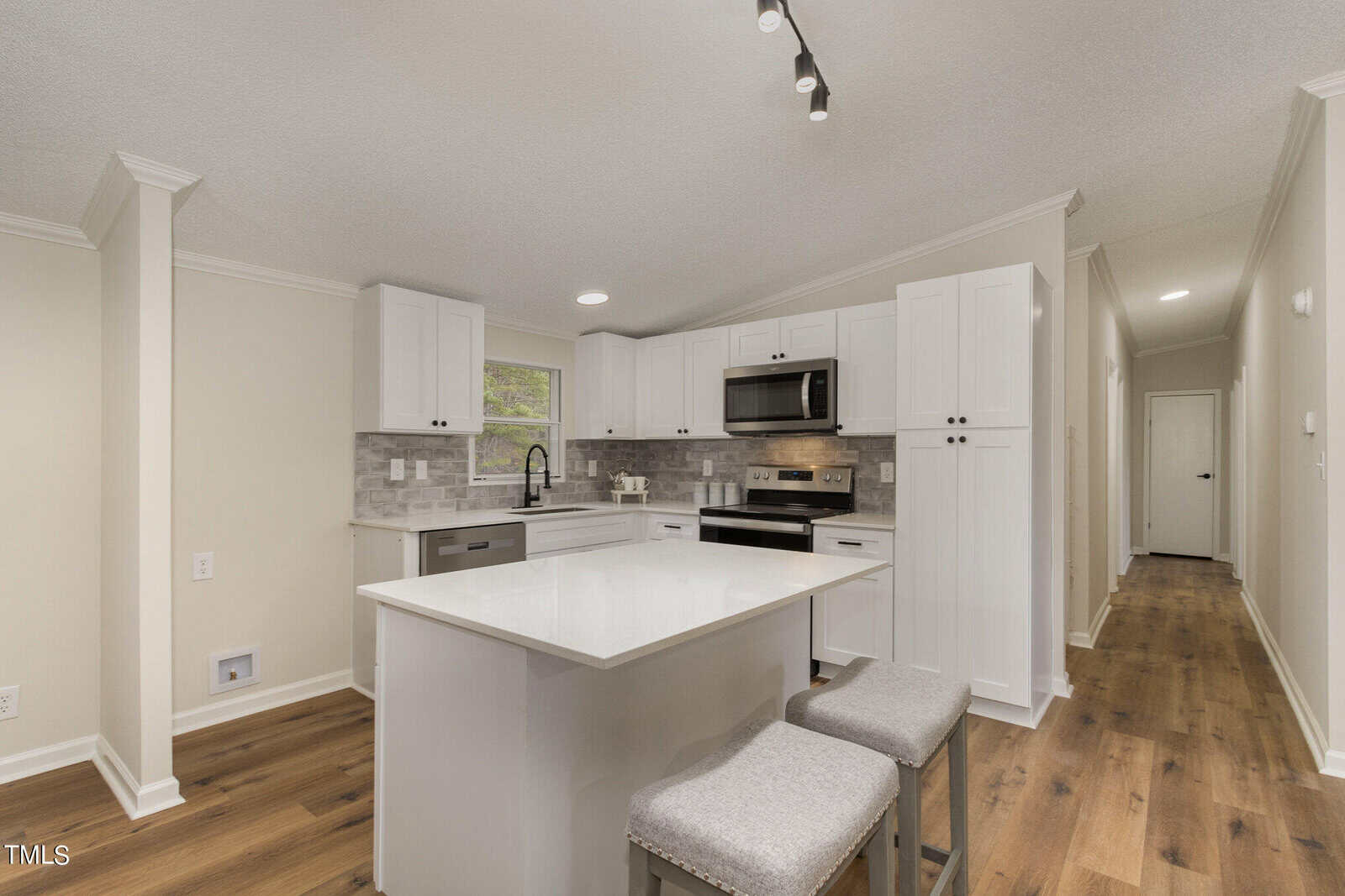 A modern kitchen with white cabinets, stainless steel appliances, and a gray backsplash. A white island with two gray cushioned stools is centered. Warm wood flooring and recessed lighting complete the space.