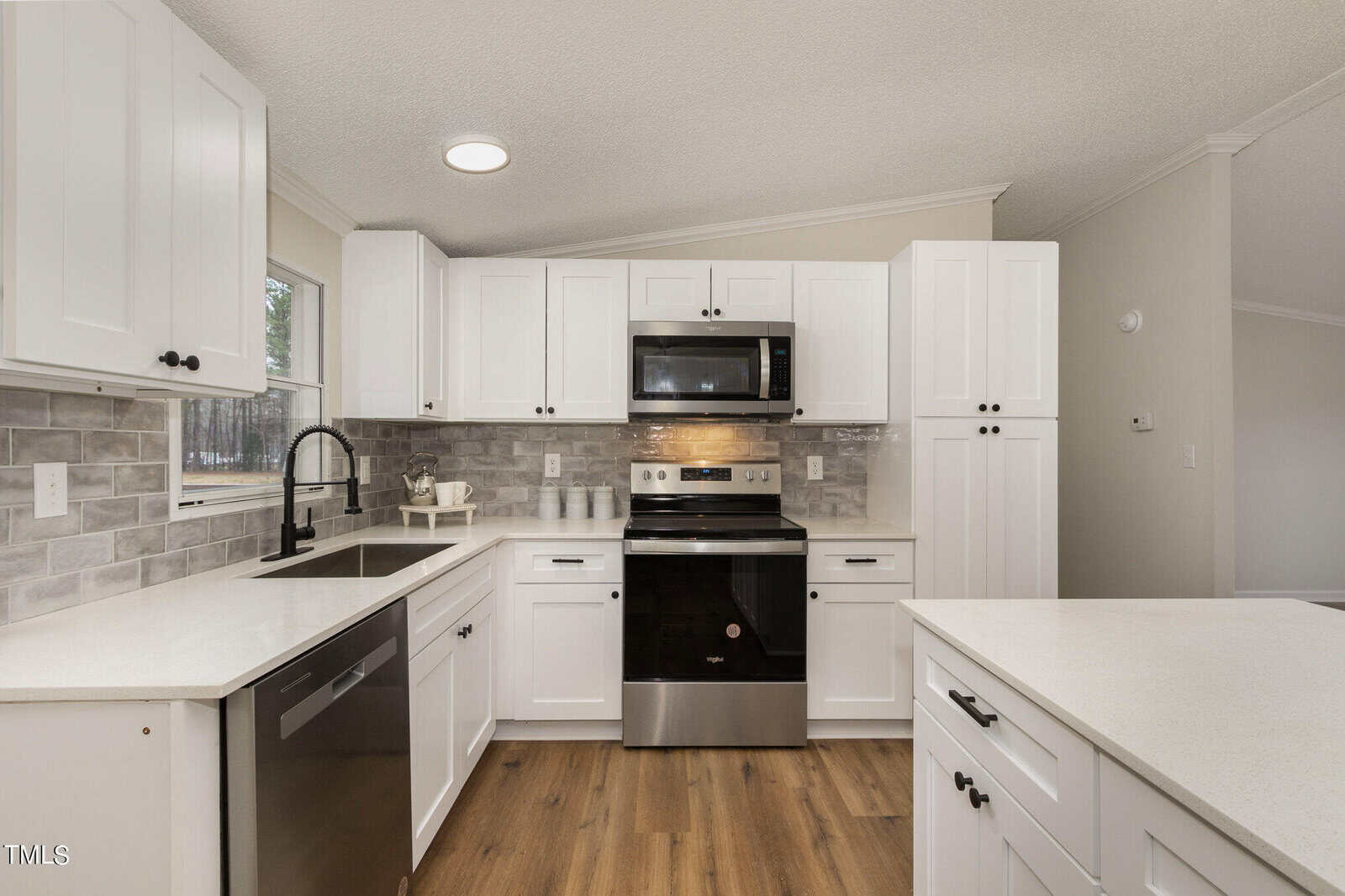 Modern kitchen with white cabinets, black appliances, and a gray tile backsplash. Features a stainless steel sink, electric stove, microwave, and dishwasher. Wood flooring adds warmth to the sleek, clean design. Natural light from a side window.