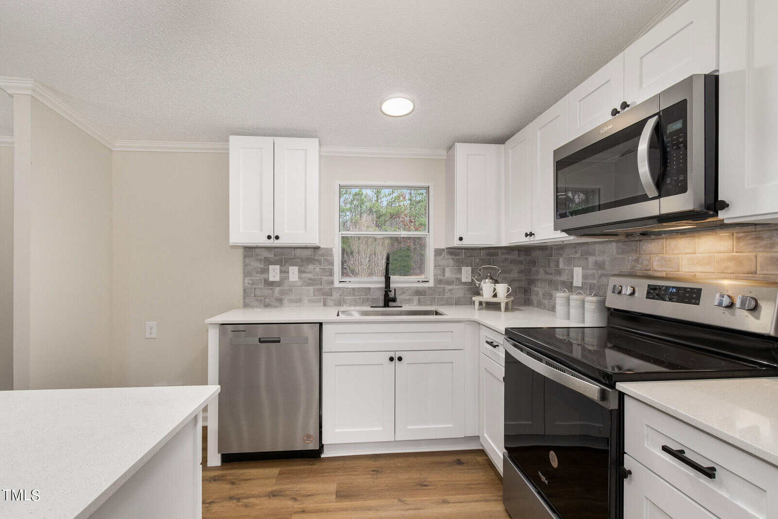 A modern kitchen with white cabinets, a stainless steel oven, and a microwave. The room features a window overlooking greenery, a gray backsplash, wooden flooring, and a sleek countertop with a built-in dishwasher.